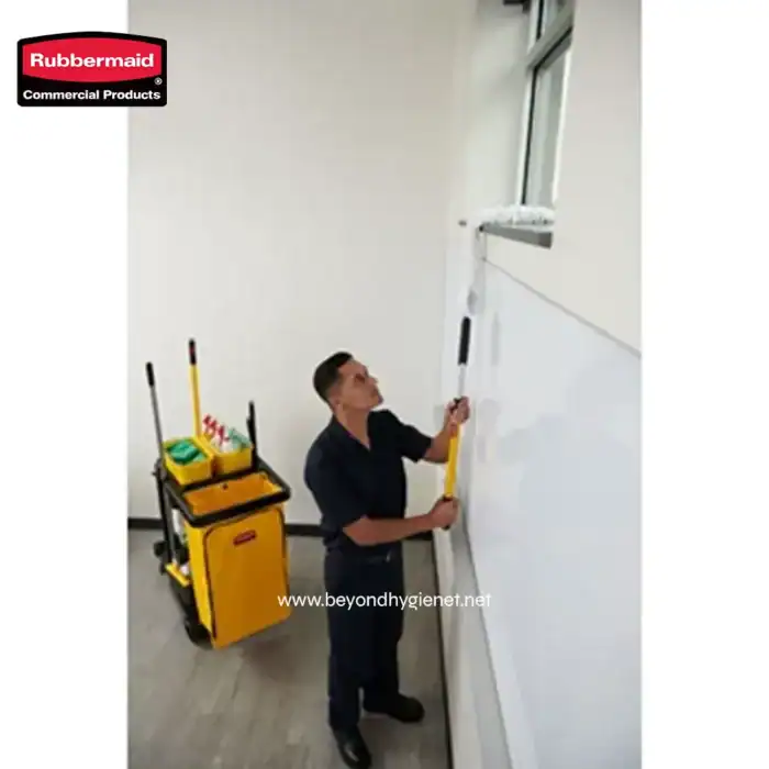 Rubbermaid commercial cleaning cart with mops and cleaning supplies beside a custodian sanitizing whiteboards in an office building.
