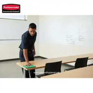 Cleaning employee disinfecting a classroom table with a green cloth, promoting hygiene and sanitation standards in educational environments.