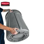 Rubbermaid commercial trash bin being held and inspected for hygiene and durability in an industrial setting. Durable, high-capacity waste receptacle ideal for commercial and sanitation needs.