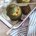 Zucchini stuffed with vegetables on a white tray, with cutlery and a blue-striped cloth, highlighting food presentation and kitchen tools.
