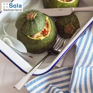 Zucchini stuffed with vegetables on a white tray, with cutlery and a blue-striped cloth, highlighting food presentation and kitchen tools.