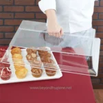 Fresh baked muffins on a white tray protected by a clear plastic cover for hygiene, at Beyond Hygiene Asia, promoting safe bakery display solutions and food safety standards.