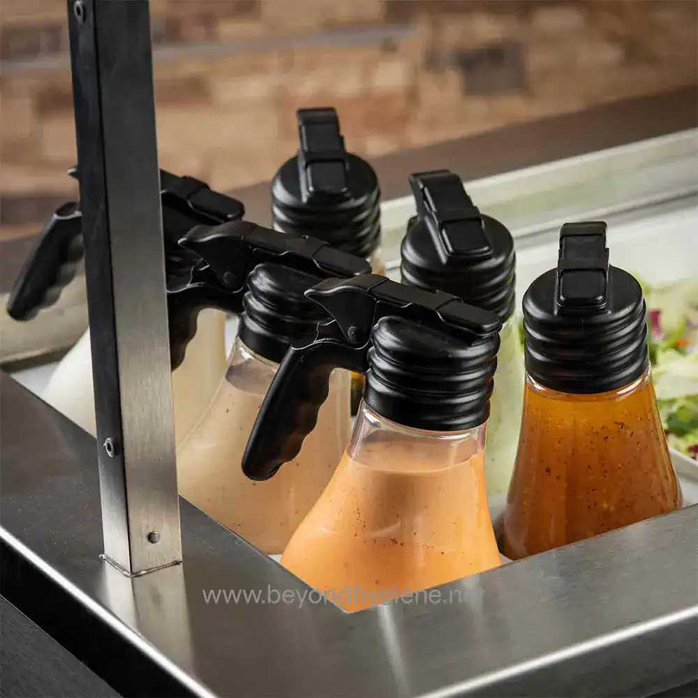 2 Qué: Bottled sauces with black spray caps in a commercial kitchen or food service setting.