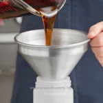 Pouring dark syrup into a metal funnel over a white container, with the context of hygiene and sanitation in food processing or beverage manufacturing.
