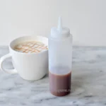 Squeeze bottle with chocolate syrup next to a coffee mug with caramel drizzle, on a marble surface, focused on food service and hygiene standards at Beyond Hygiene Asia.