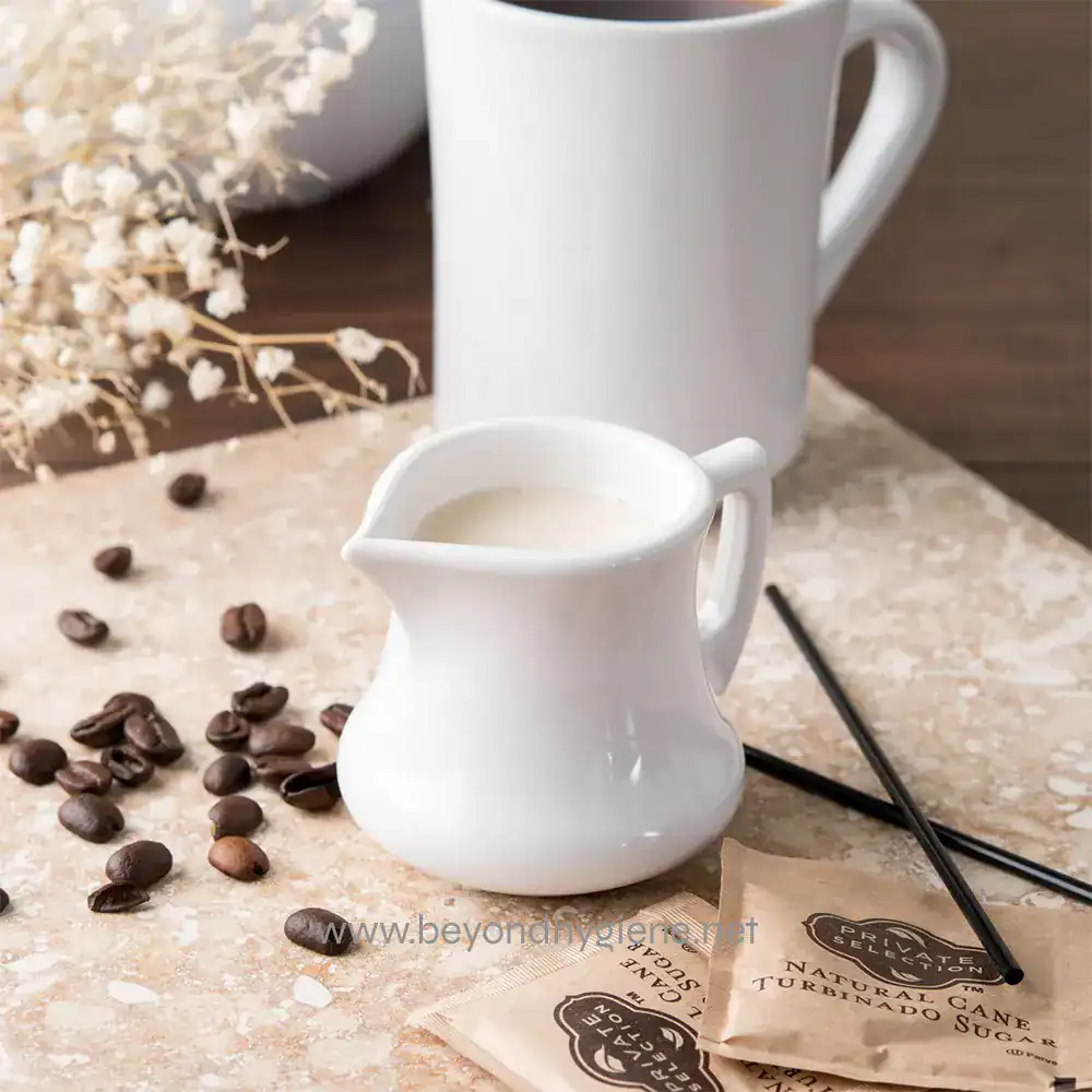 Creamy milk served in a small white pitcher with coffee beans scattered around on a textured surface, with a white mug and dried flowers in the background.