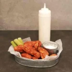 Crispy buffalo chicken wings with celery sticks and creamy dipping sauce on a metal tray, with a squeeze bottle of ranch or blue cheese dressing in the background.