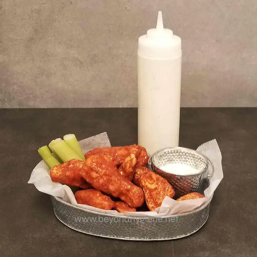 6 Crispy buffalo chicken wings with celery sticks and creamy dipping sauce on a metal tray, with a squeeze bottle of ranch or blue cheese dressing in the background.