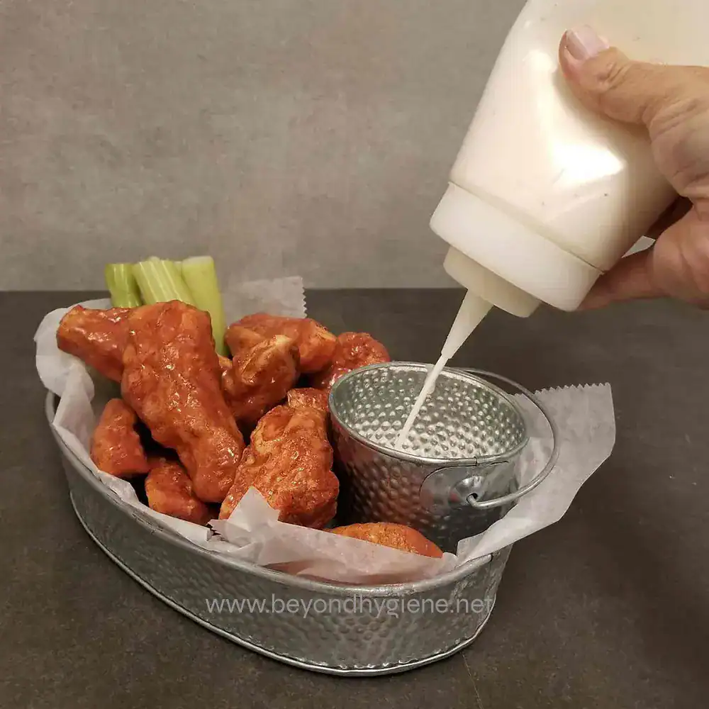 7 Crispy buffalo chicken wings with celery sticks and ranch sauce, being topped with creamy dressing, served in a metal basket, perfect for food presentation and snack or appetizer, beyond hygiene asia.