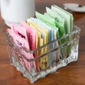 Dispenser of colorful sugar packets in a clear glass container on a wooden table at Beyond Hygiene Asia.