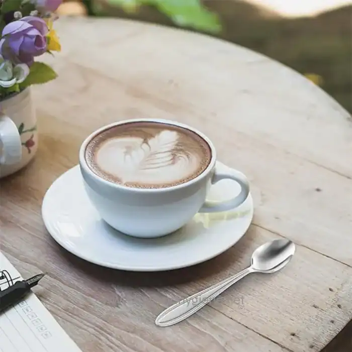 Coffee in a white cup with latte art on a wooden table, beside a spoon and a potted plant, representing hygiene and cleanliness in beverage service.