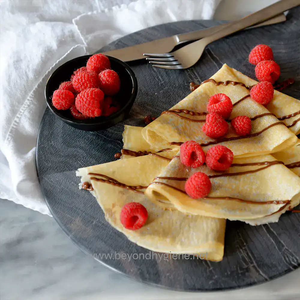 9 Fresh crepes topped with raspberries and drizzled with chocolate on a dark plate, served with a side of fresh raspberries, perfect for breakfast or dessert at Beyond Hygiene Asia.