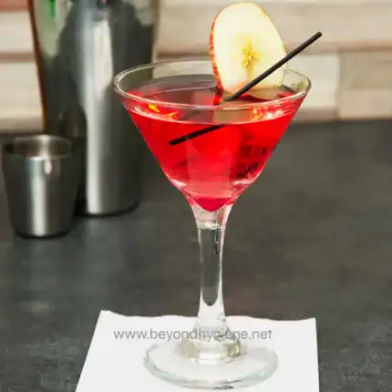 Pink cocktail with apple slice garnish in a martini glass on a bar counter, showcasing beverage and hospitality services.