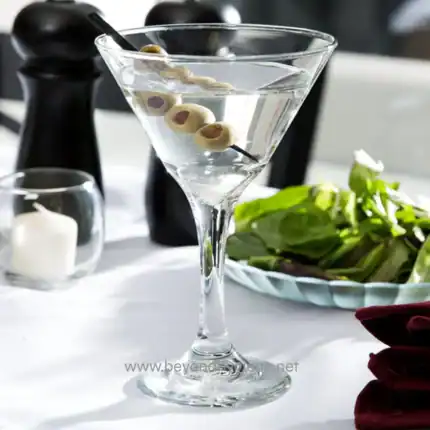Olive martini with green olives in a clear cocktail glass, served on a white table with greenery and candles in the background.