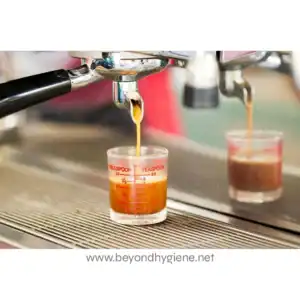 Measuring espresso shot from commercial espresso machine in a glass cup, professional coffee brewing process, hot espresso being poured from portafilter spout, Beyond Hygiene Asia logo at the bottom.