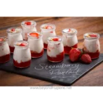 Strawberry parfait desserts in small glass jars, creamy yogurt layered with fresh strawberries and strawberry sauce, served on a rustic wooden table with whole strawberries nearby.