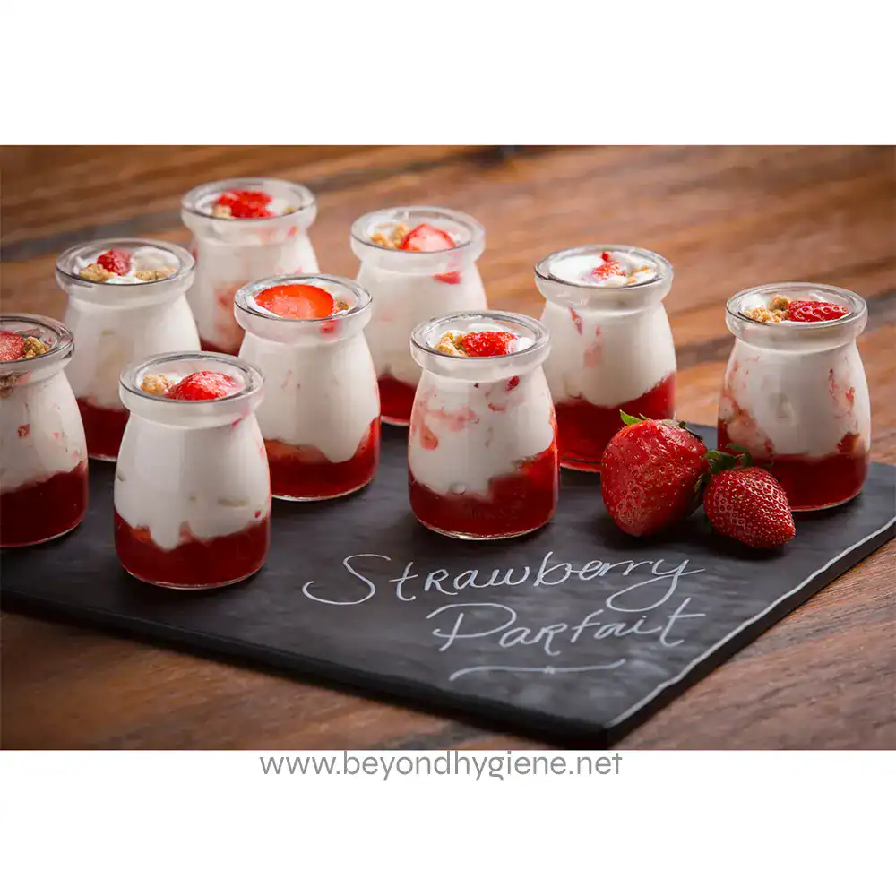 MG1_ENV-01 Strawberry parfait desserts in small glass jars, creamy yogurt layered with fresh strawberries and strawberry sauce, served on a rustic wooden table with whole strawberries nearby.
