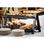 Freshly baked cupcakes, cookies, and pastries displayed on a black two-tier food stand at a catering event, emphasizing food safety and hygiene.