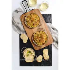 Spiced Indian biryani with fried chicken, served with hummus and naan bread, accompanied by two glasses of white wine on a stylish presentation tray.
