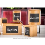 Wooden storage boxes with chalkboard labels for kitchen organization, featuring categories like sugar packs, napkins, straws, and silverware, set in a modern kitchen.